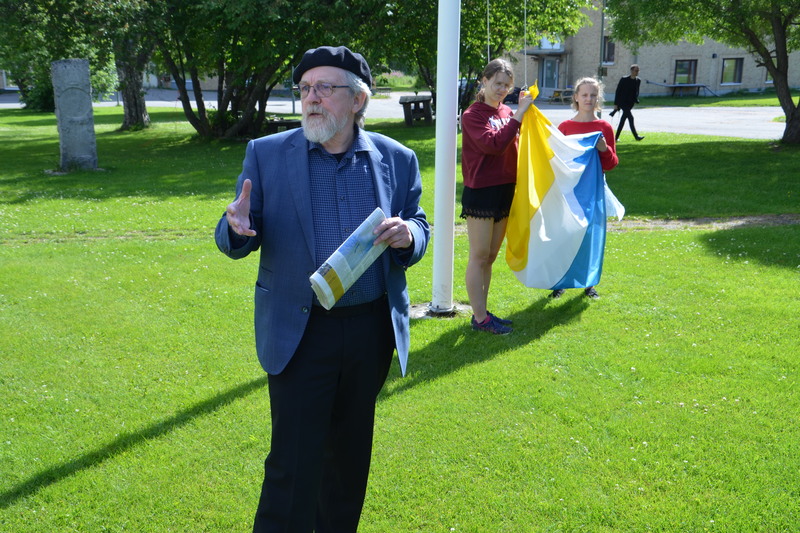 En man i basker står framför två andra personer som är i färd att hissa en flagga med färgerna vitt, gult och blått.