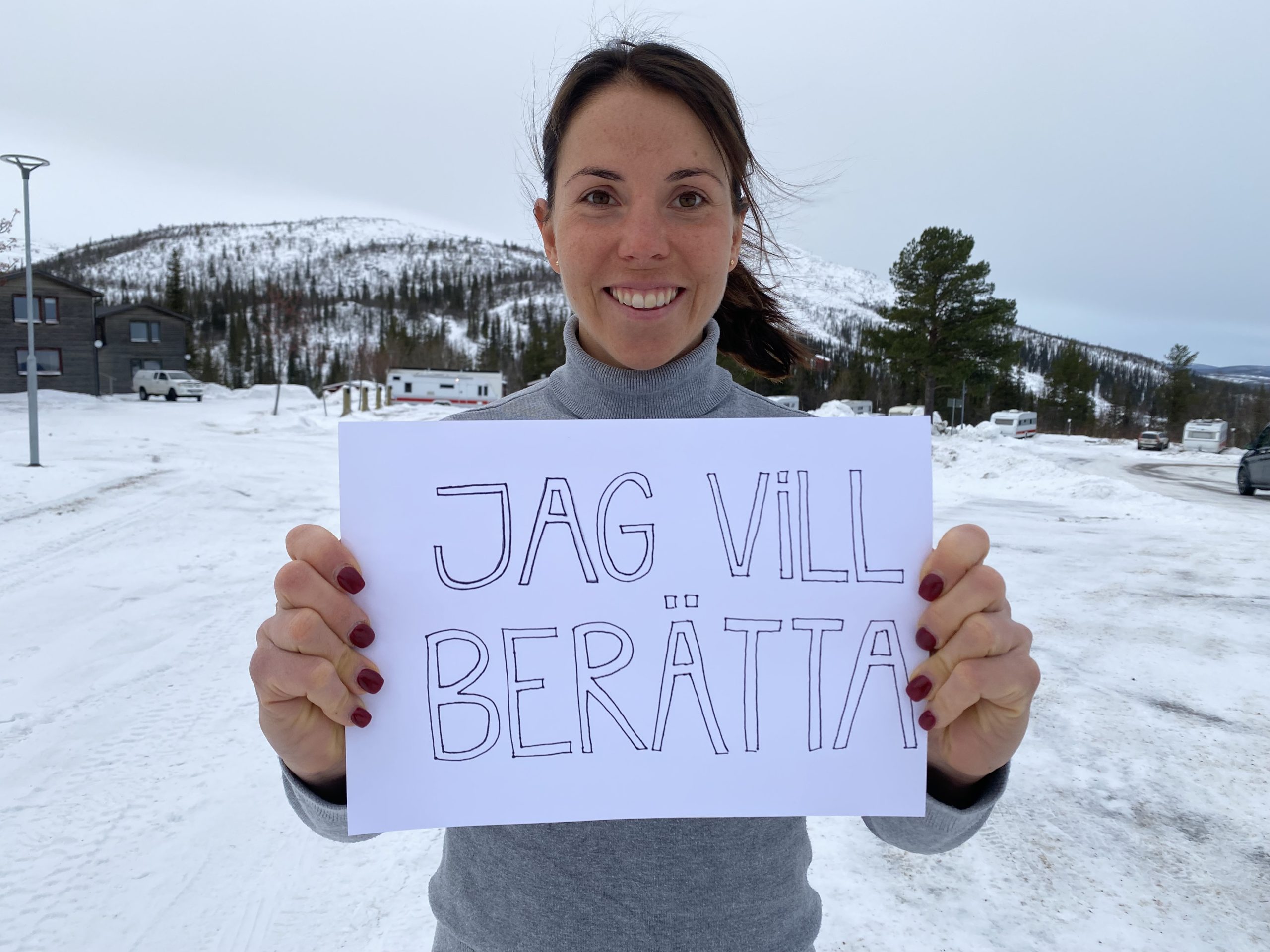 En kvinna står leende i ett snölandskap och håller upp en skylt. På skylten står "jag vill berätta".