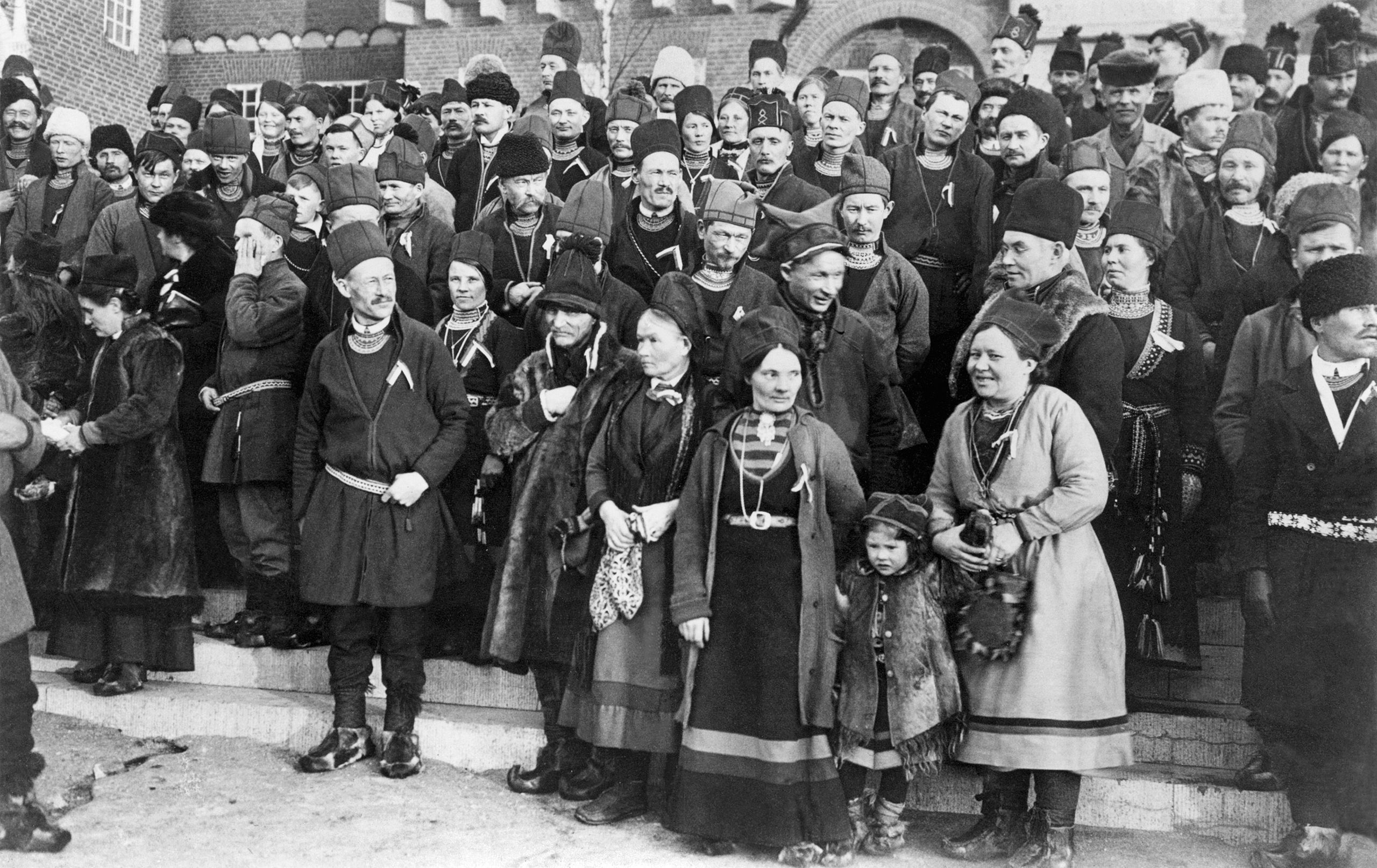 Svartvitt foto av ett 40-tal människor i samiska kläder som står uppställda för gruppfoto på en trappa.