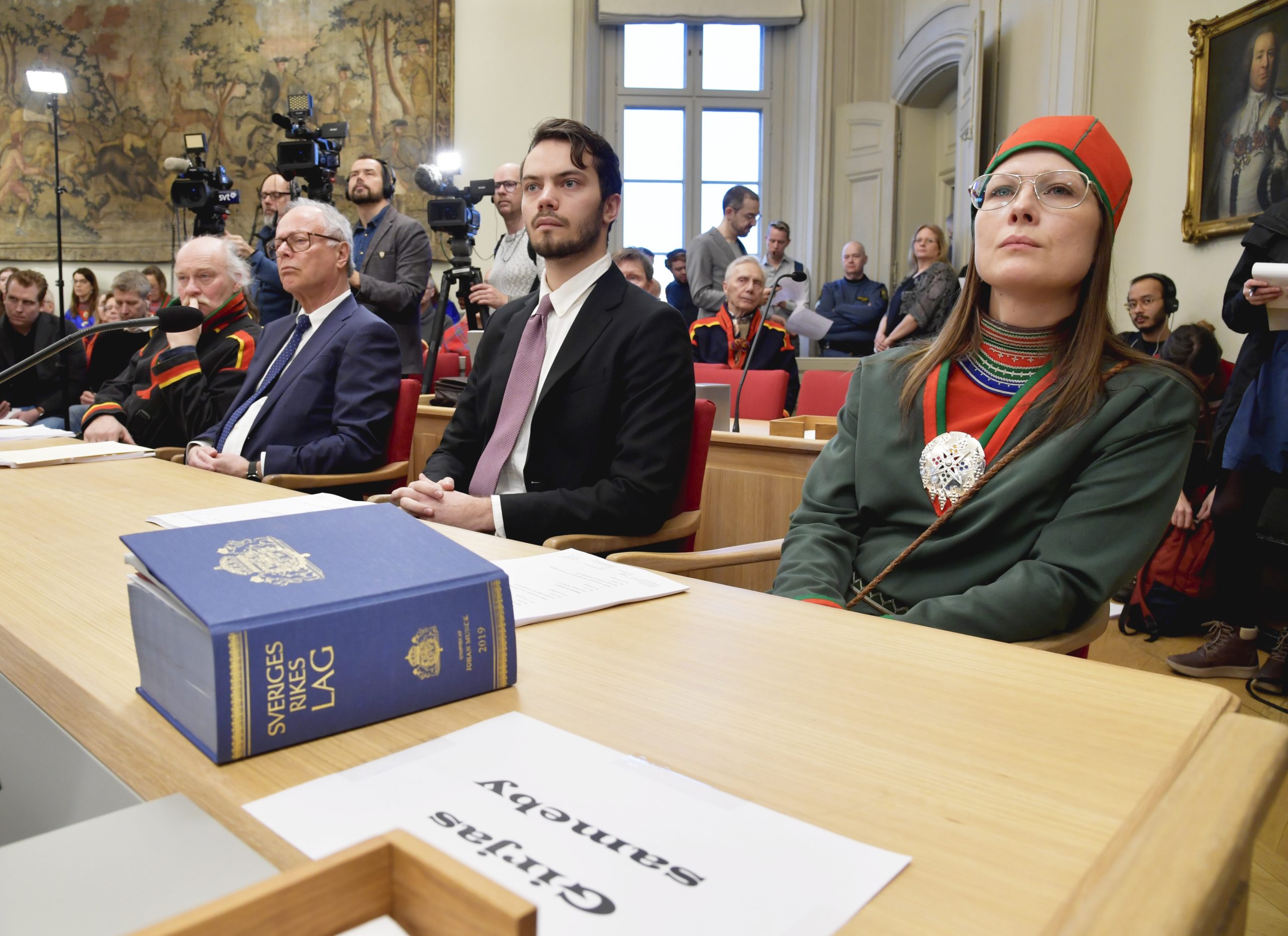 Personer sitter uppradade framför en mikrofon med en lagbok framför sig. Några av personerna bär kläder som skulle kunna vara traditionellt samiska och andra kostymer.