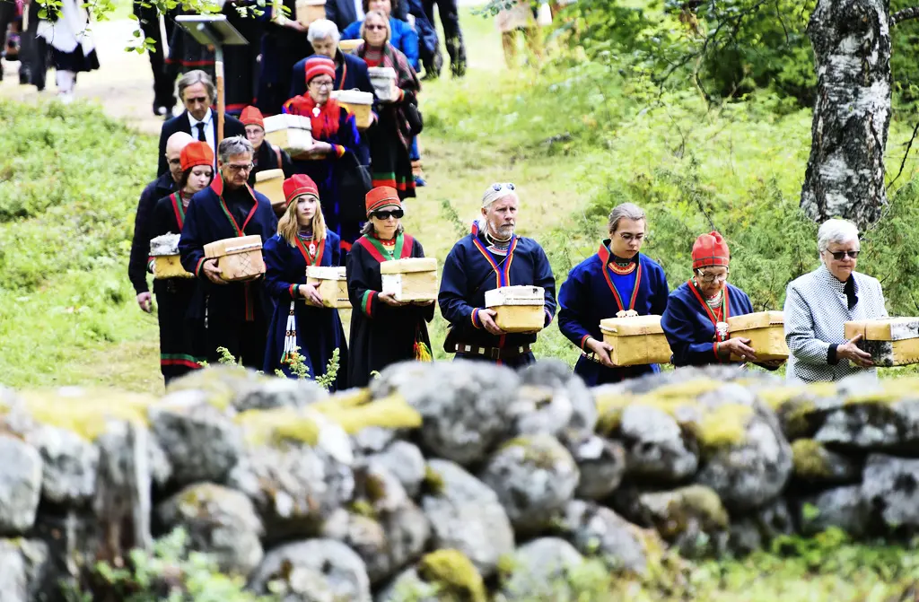 En procession av människor i kläder med samiskt röda och blå färger. Människorna bär på askar av näver.