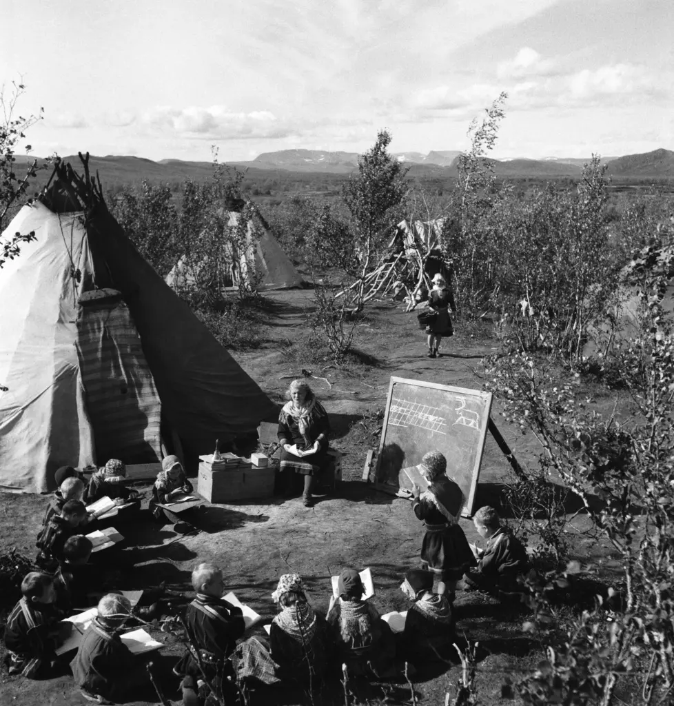 Svartvitt foto. Framför ett tält i fjällmiljö sitter barn i ring framför en svart griffeltavla.